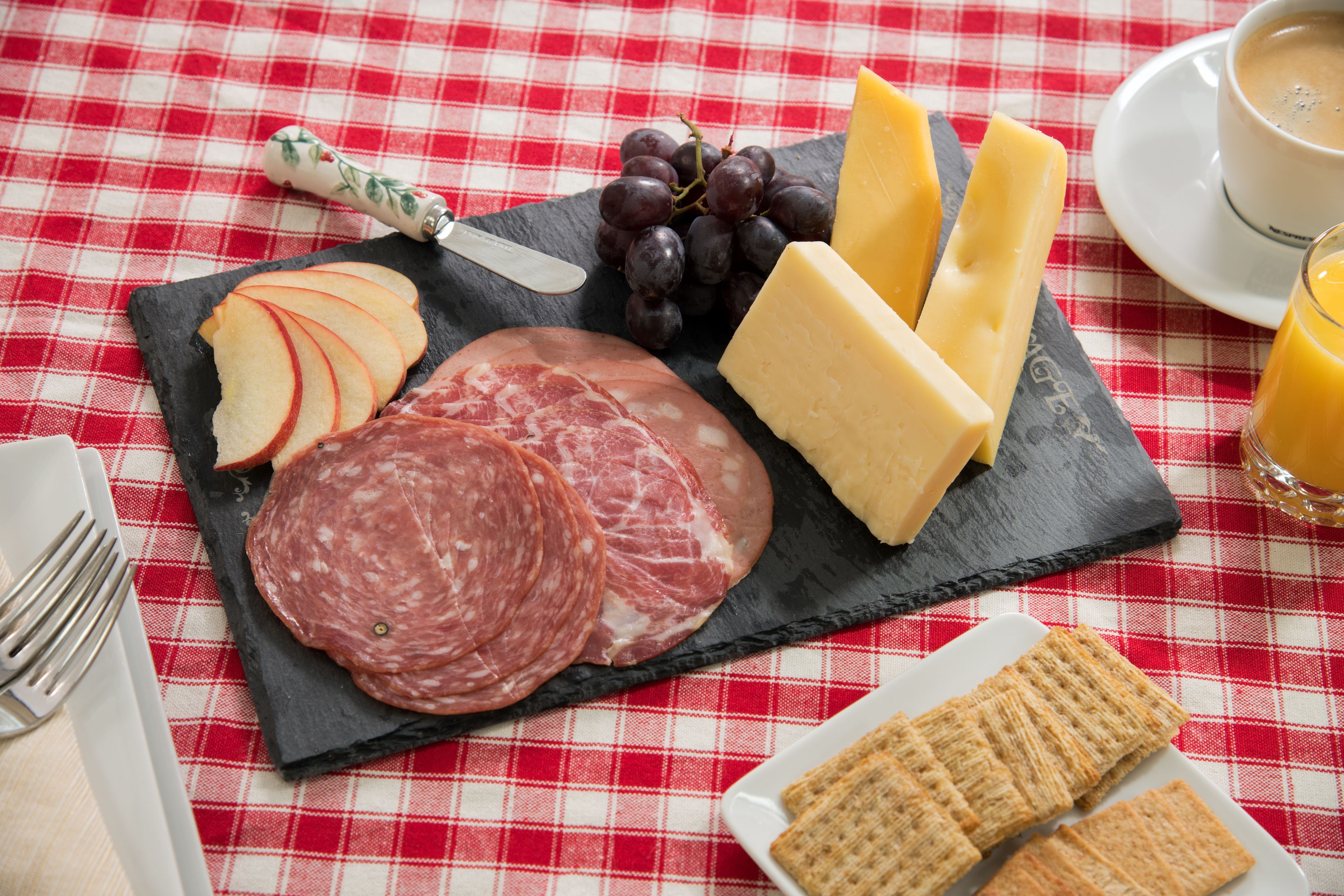 A meal setup with sliced apples, cured meats, cheese, and crackers on a slate board, accompanied by a cup of coffee and a glass of orange juice on a red and white checkered tablecloth.