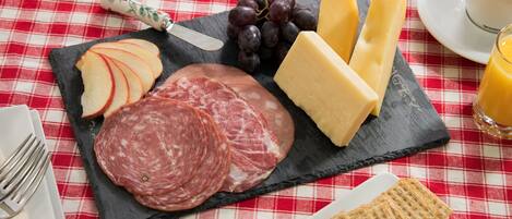A meal setup with sliced apples, cured meats, cheese, and crackers on a slate board, accompanied by a cup of coffee and a glass of orange juice on a red and white checkered tablecloth.