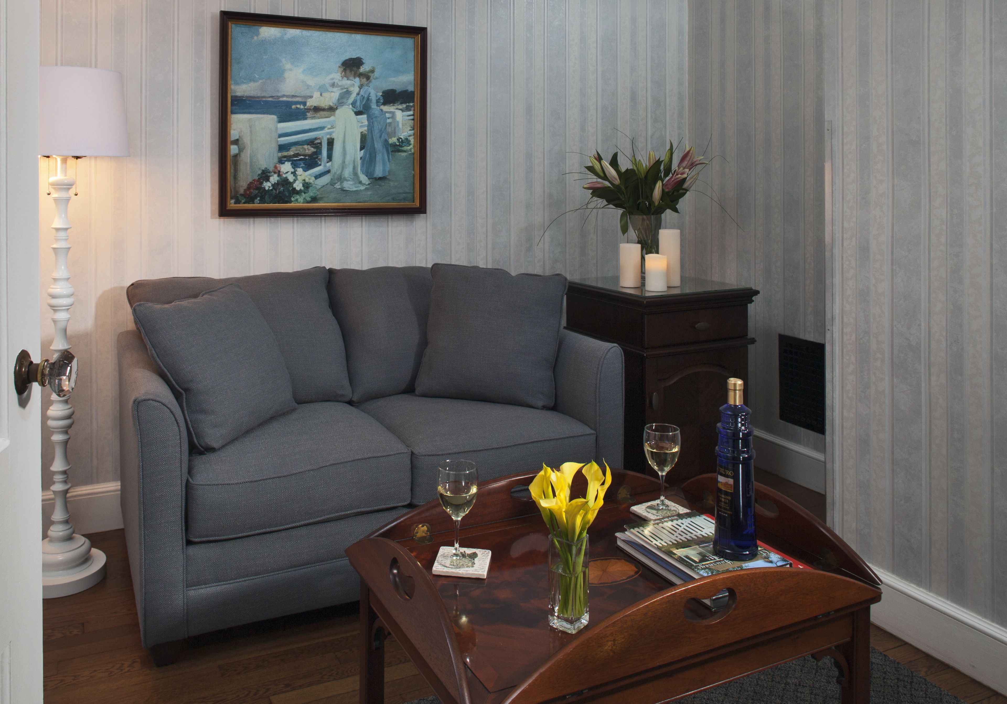 A living room with a grey sofa, a wooden coffee table with a vase of yellow flowers, a bottle of champagne, and a framed painting on the wall.