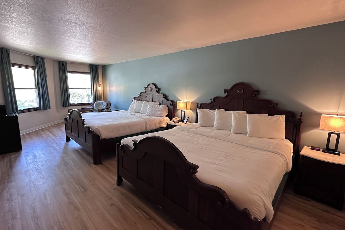 A hotel room with two wooden beds, a small desk, a chair, and a window with curtains.