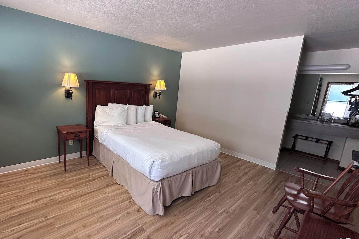 A hotel room with a bed, two bedside lamps, a nightstand, a wooden floor, and a small desk with a chair.