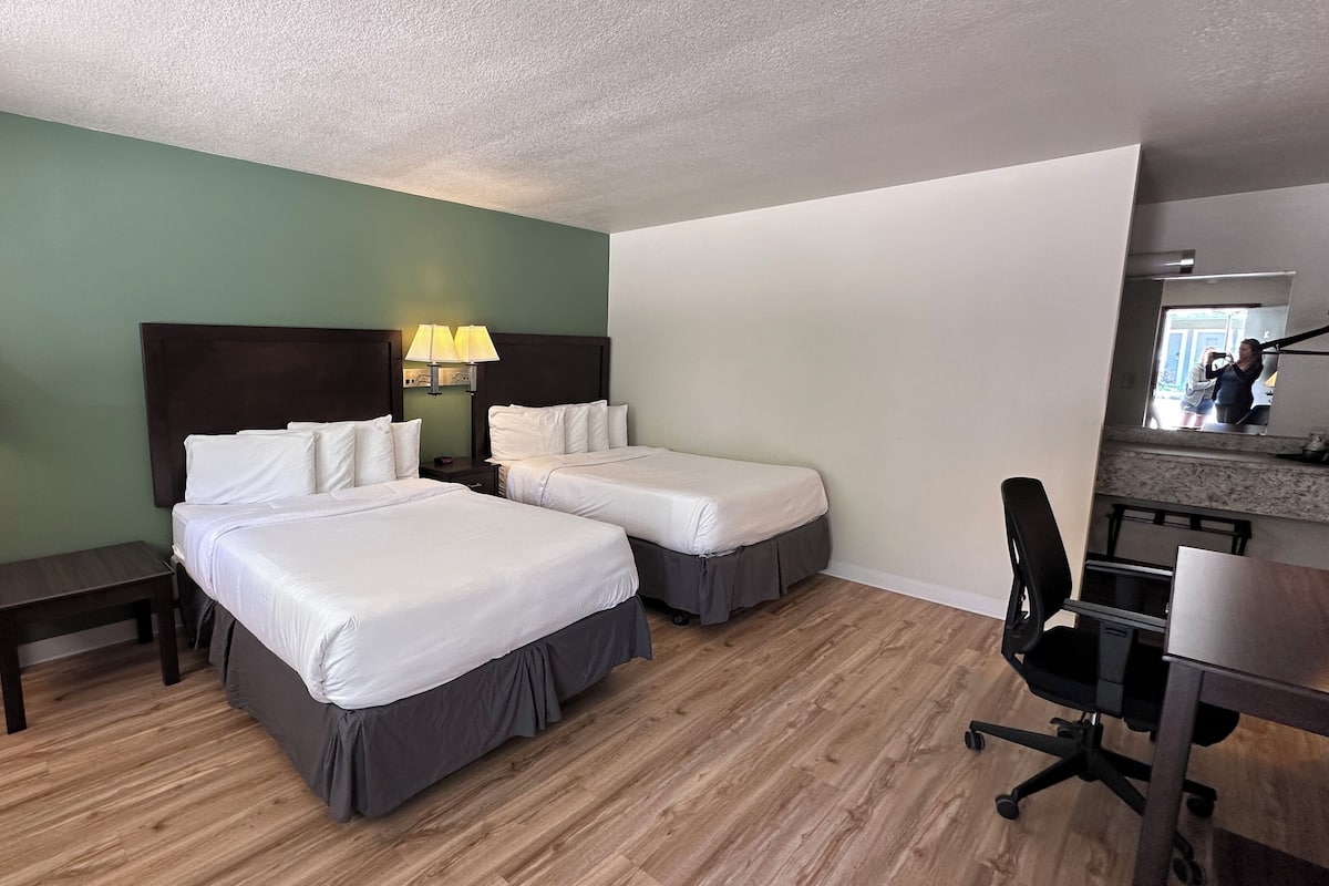 A hotel room with two beds, a desk, a chair, a lamp, and a mirror.