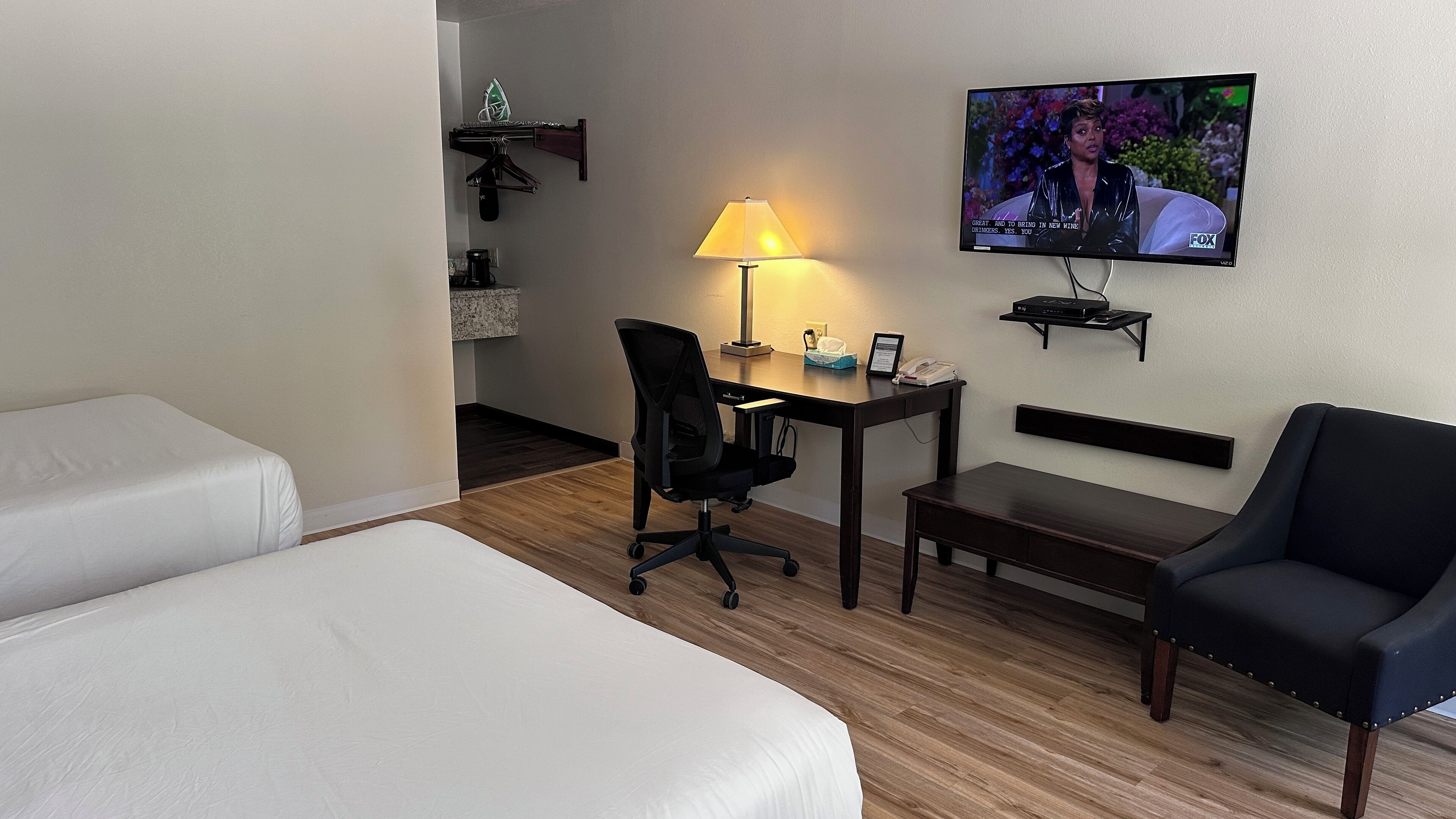 A hotel room with two single beds, a desk with a lamp, a chair, a TV mounted on the wall, and a small shelf with a coffee maker and other items.