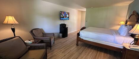 A hotel room with a bed, a chair, a small table, a TV, and a desk with a lamp.