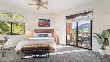 A modern hotel room with a large bed, a desk, a ceiling fan, and a balcony with a view of the mountains.