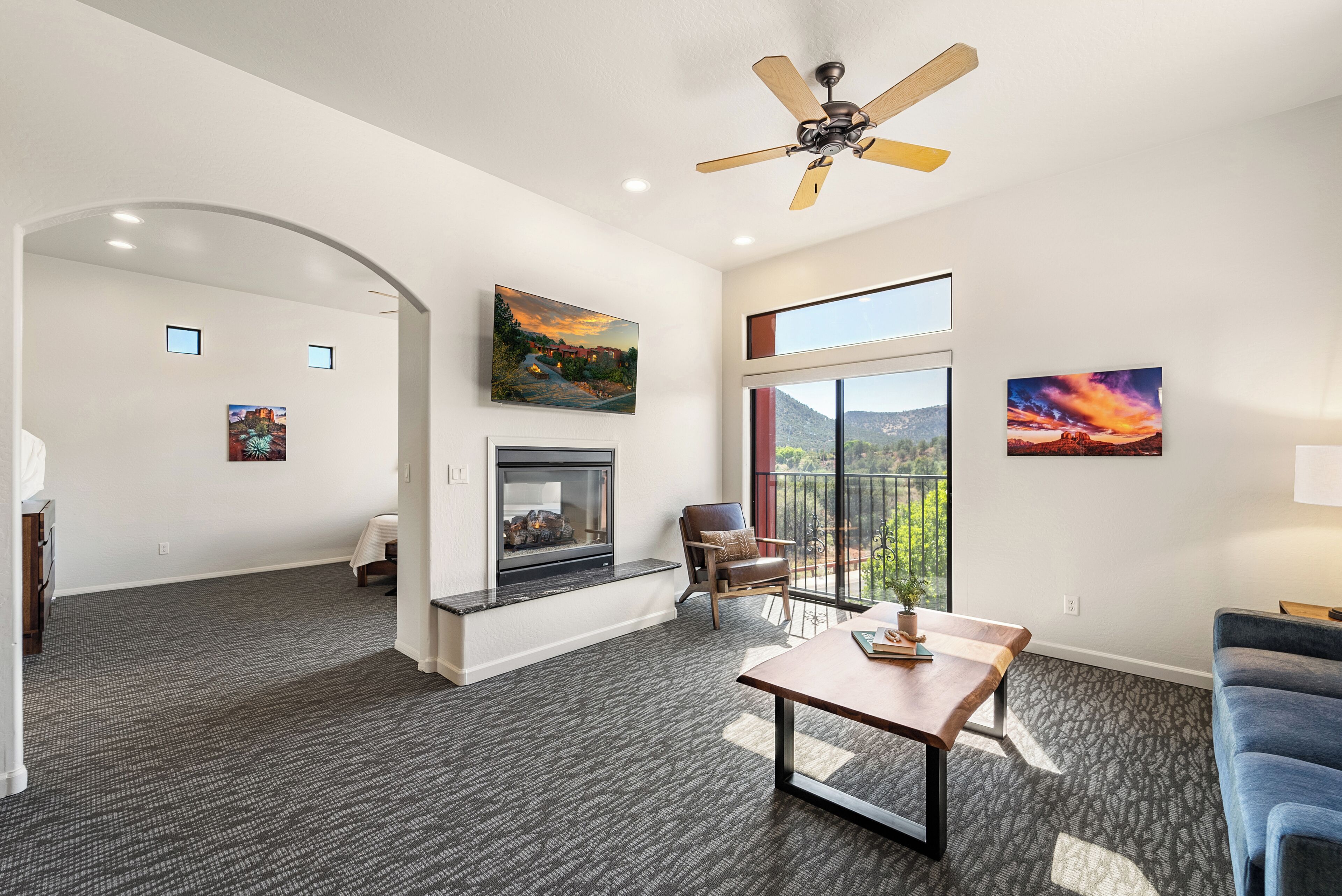 A spacious living room with a fireplace, a ceiling fan, and large windows offering a view of the outdoors.