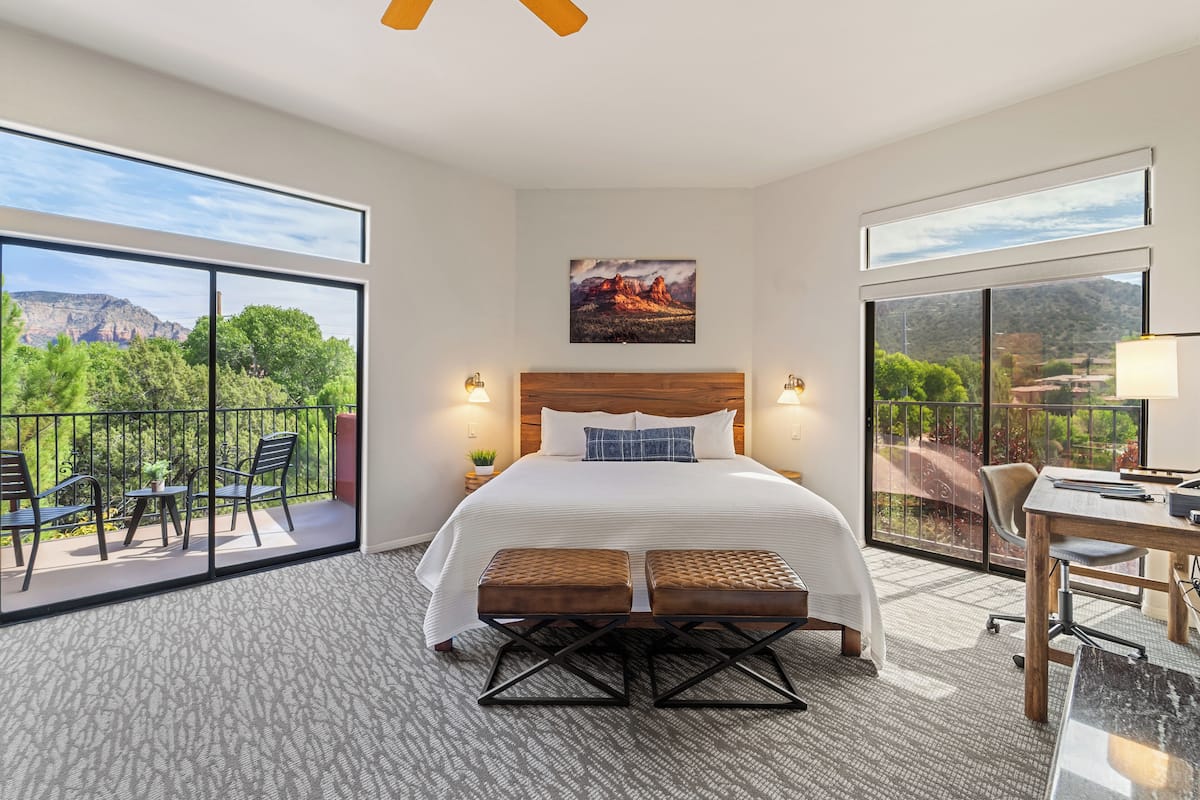 Habitación de hotel con una cama grande, un balcón con sillas, un escritorio y una vista panorámica a la montaña.