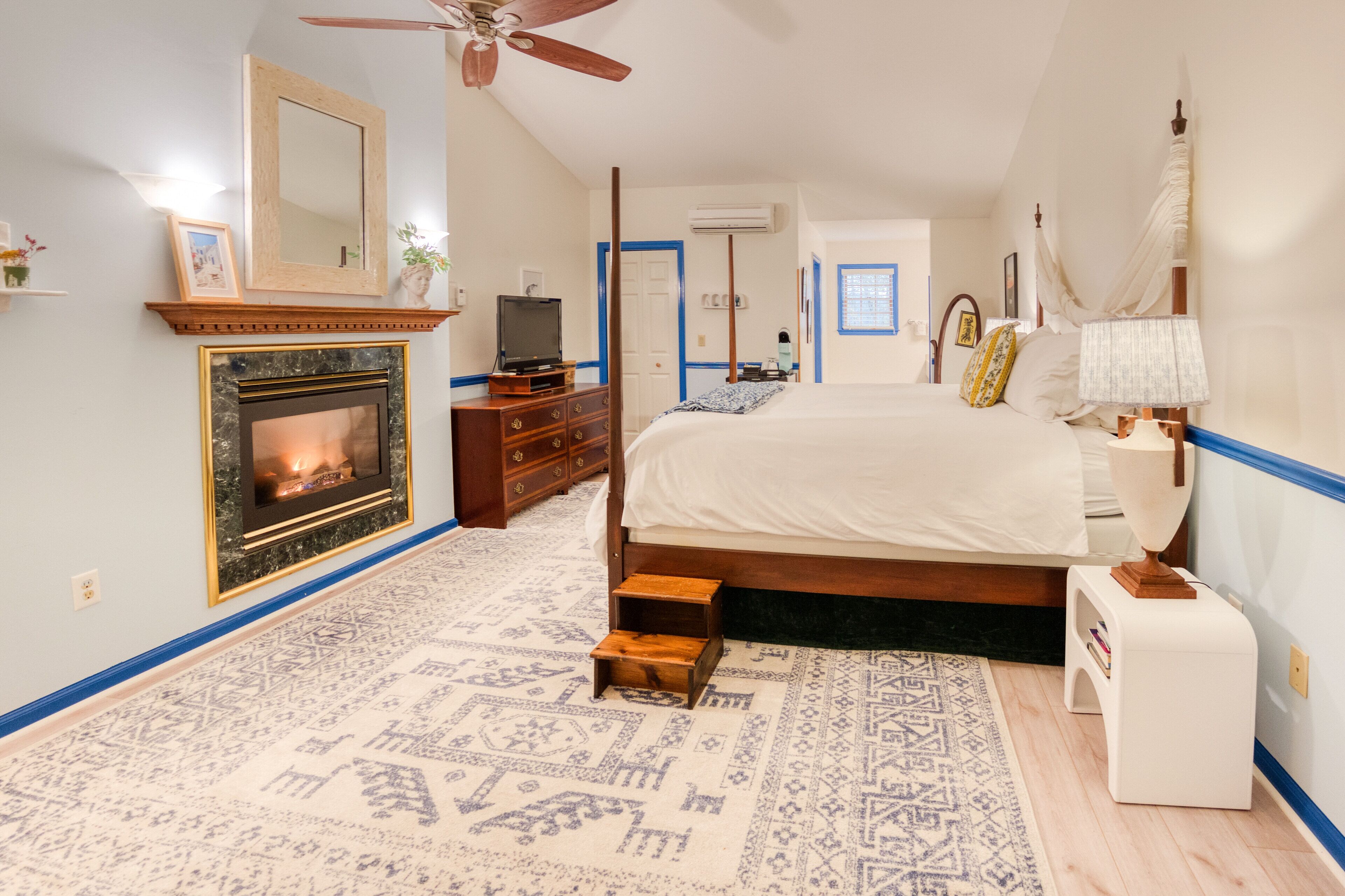 Une chambre à coucher avec une cheminée, un lit, une table de chevet, une petite table, un ventilateur de plafond et une fenêtre.