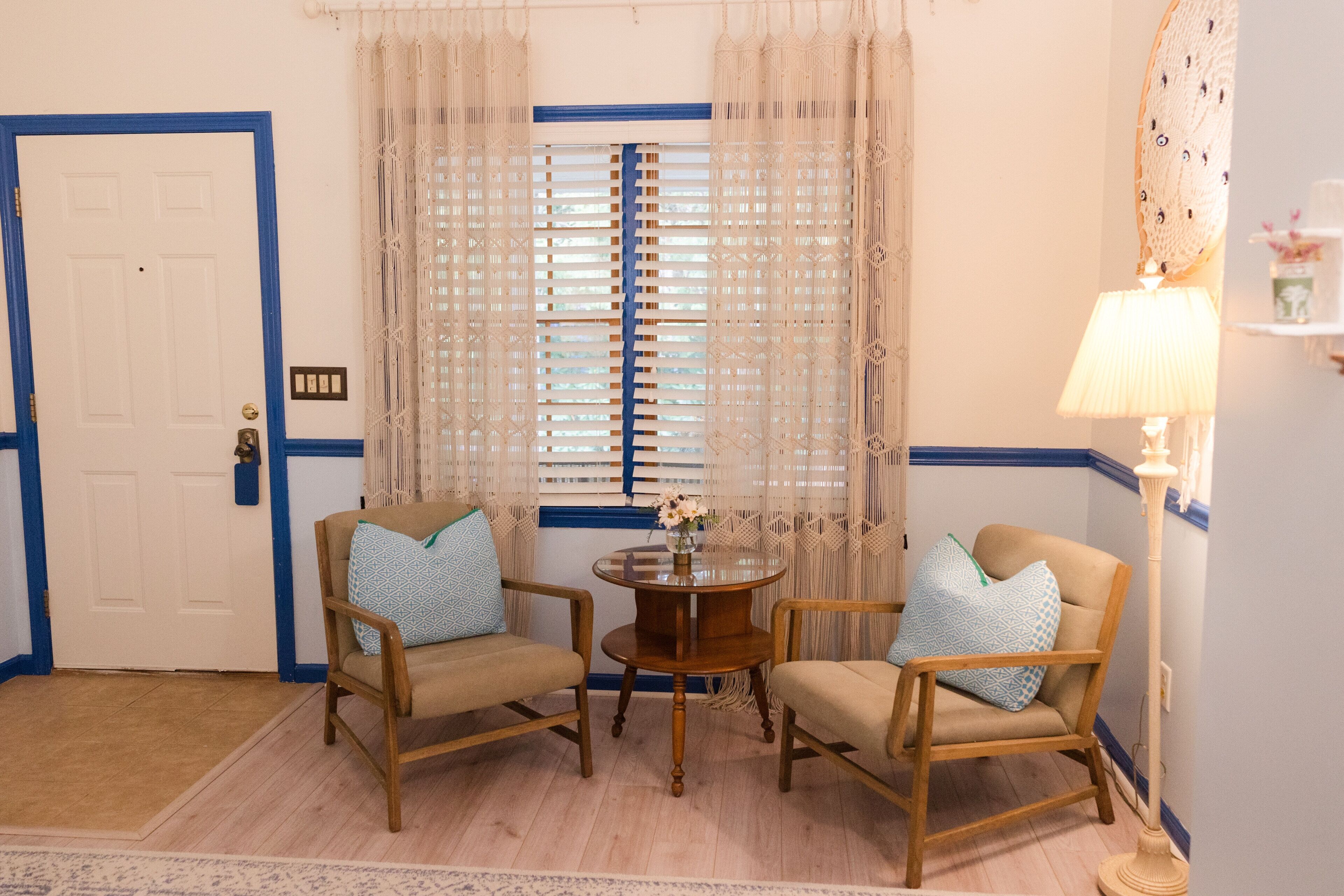 Una habitación con dos sillas de madera, una pequeña mesa redonda con un florero, una puerta con marco azul y una ventana con cortinas estampadas.