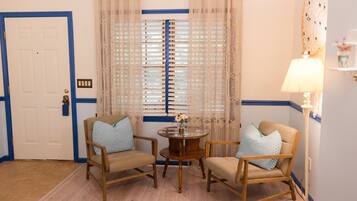 Una habitación con dos sillas de madera, una pequeña mesa redonda con un florero, una puerta con marco azul y una ventana con cortinas estampadas.