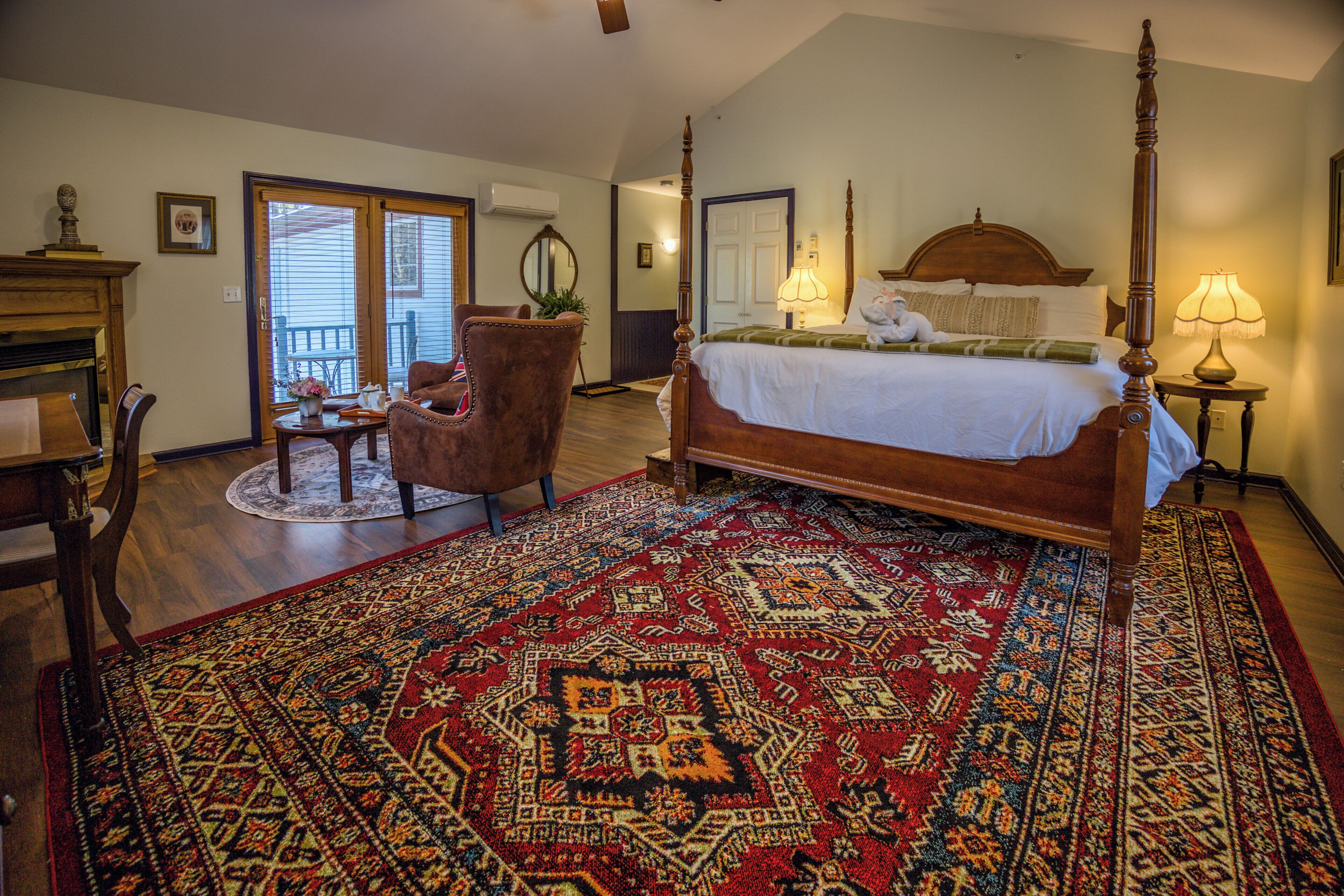 A bedroom with a four-poster bed, a large patterned rug, a chair, and a small table with a vase.