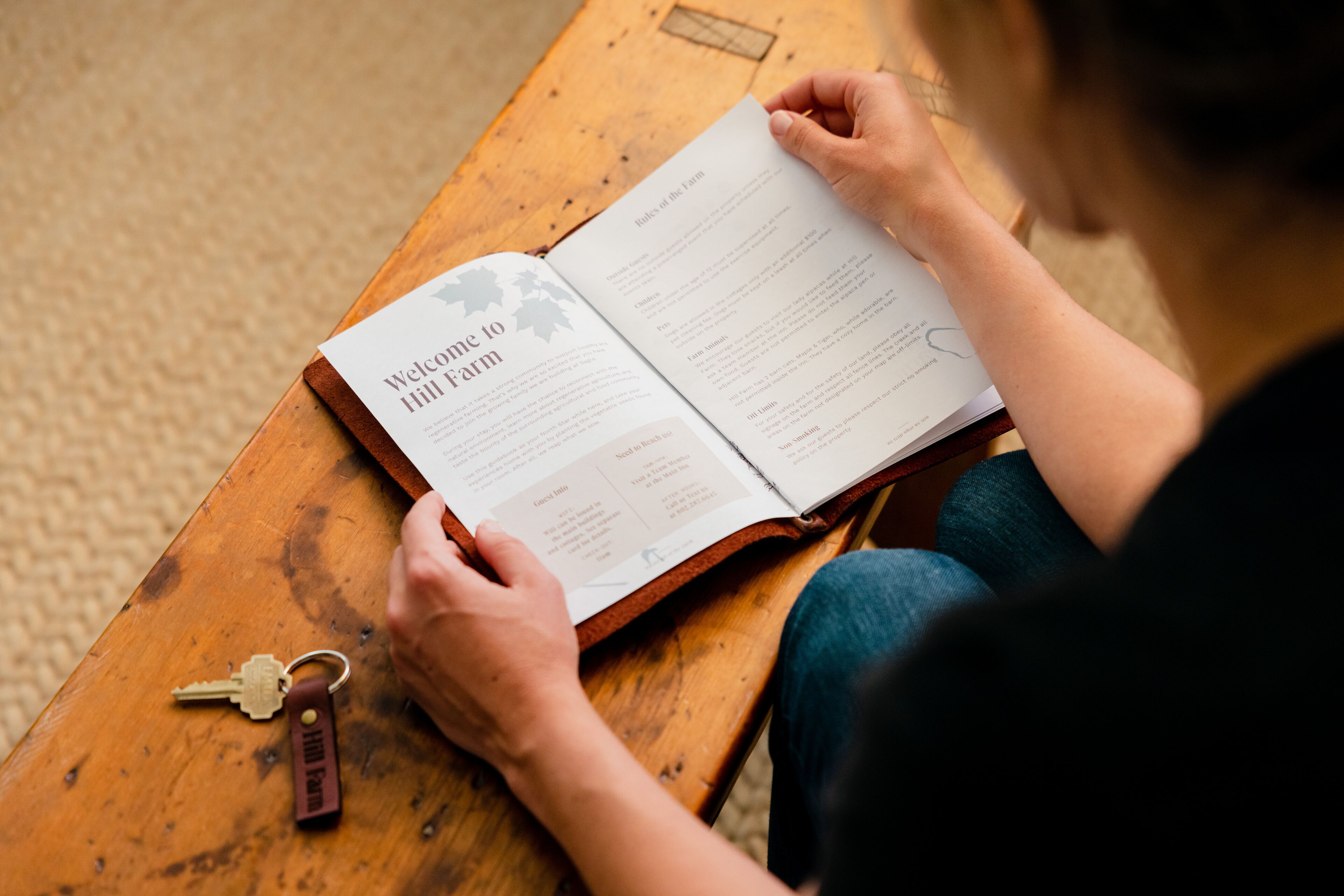 Una persona leyendo un libro titulado “Welcome to Hill Farm” sobre una mesa de madera.