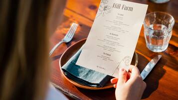 Un menú con el título "Hill Farm" sobre una mesa de madera, junto a un vaso de agua y cubiertos.