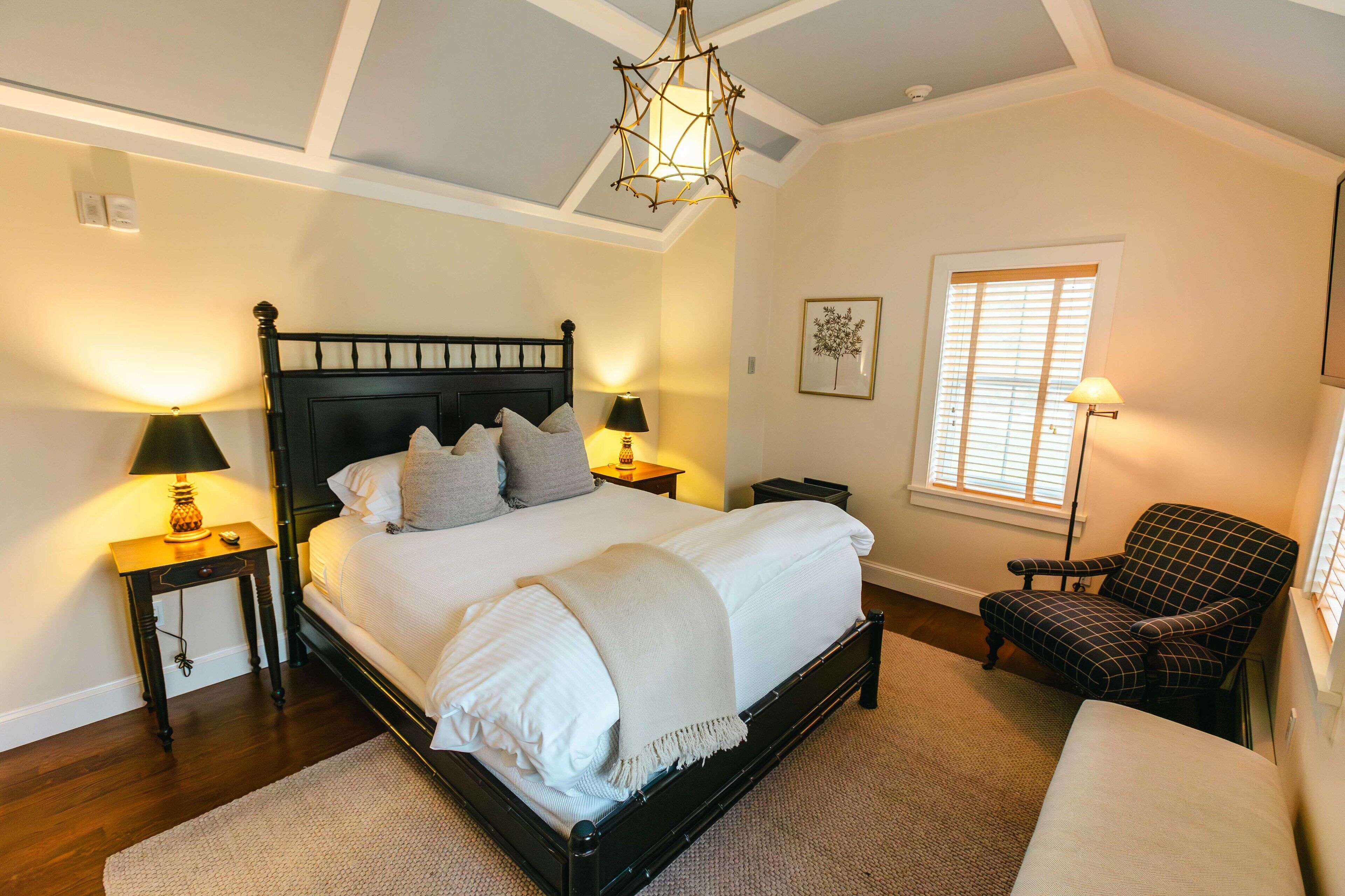 A bedroom with a large bed, two bedside tables with lamps, a chair, and a window with blinds.