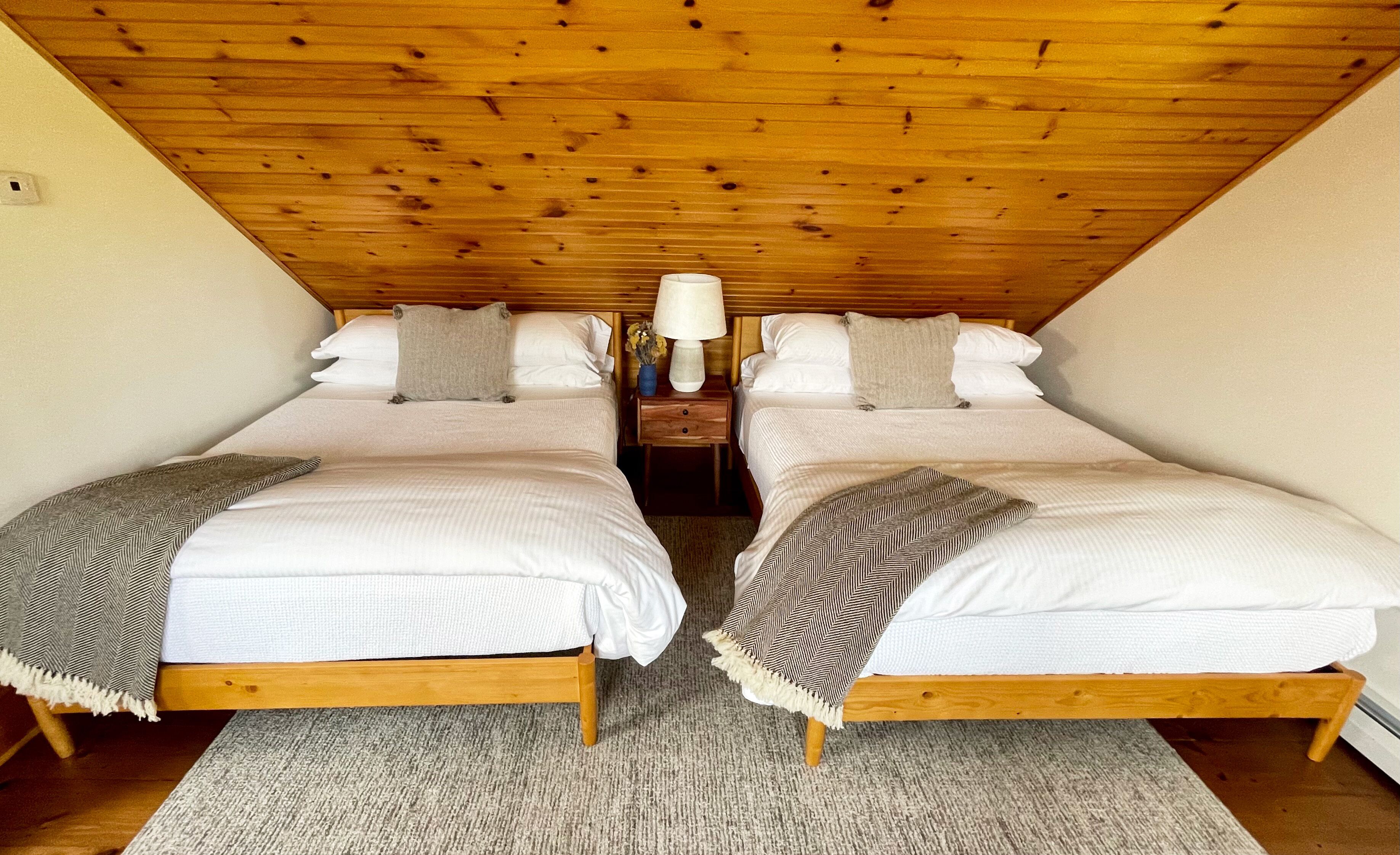Una habitación con dos camas individuales, techo de madera, una mesita de noche con lámpara y una alfombra.