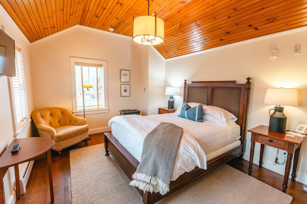 A bedroom with a wooden ceiling, a large bed with white bedding, a bedside table with a lamp, a chair, and a window with blinds.