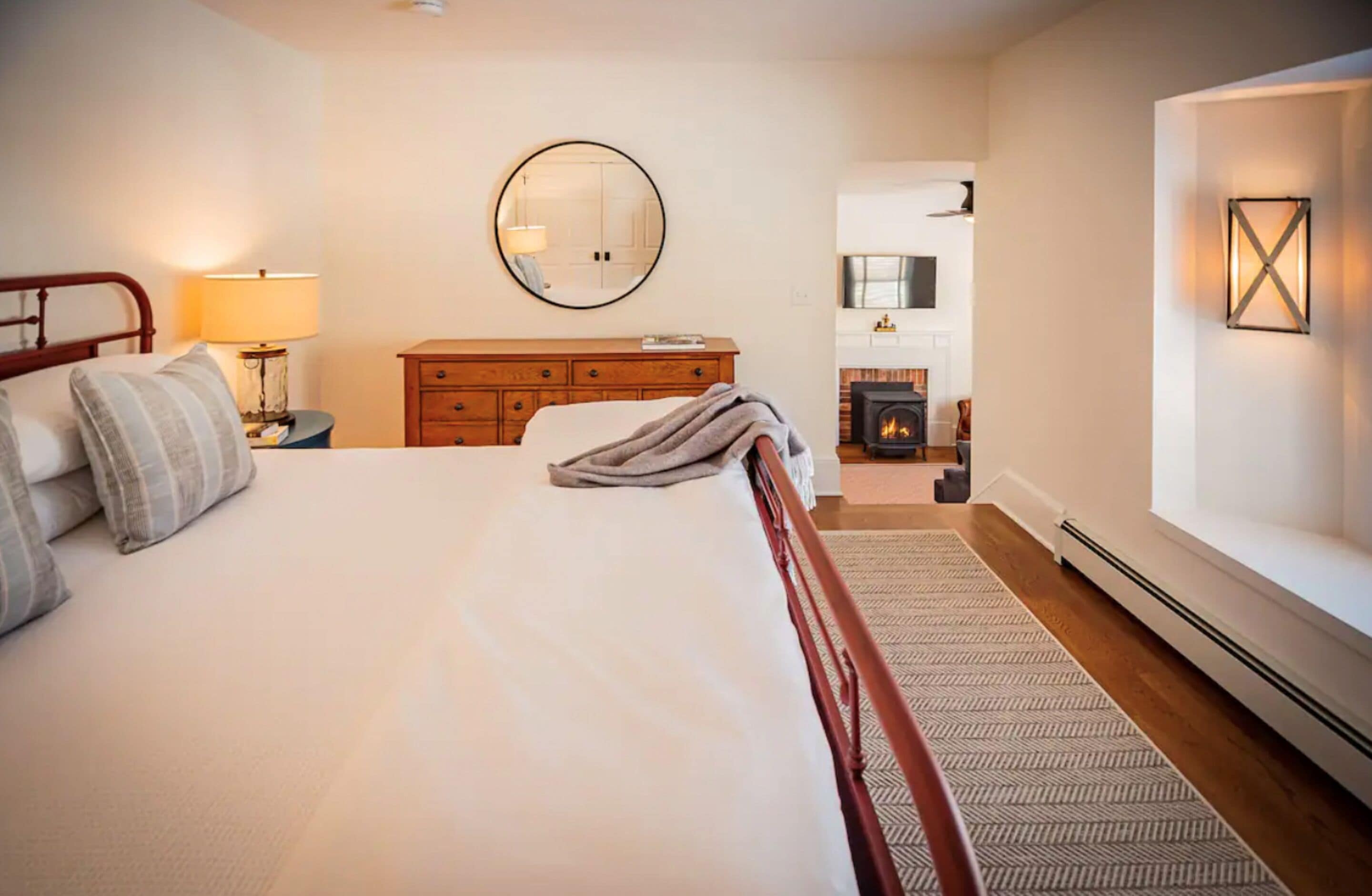 A bedroom with a large bed, a dresser, a round mirror, a fireplace, and a wall-mounted light fixture.