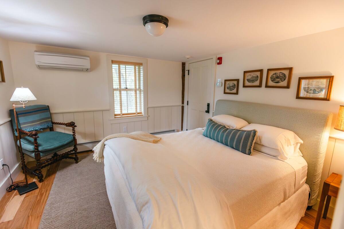 A bedroom with a large bed, a wooden chair, a window, and wall-mounted pictures.