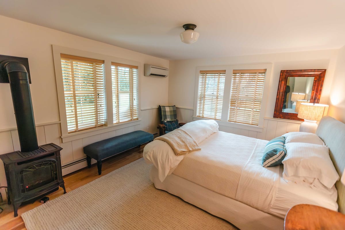 A bedroom with a large bed, a bench, a fireplace, and windows with blinds.