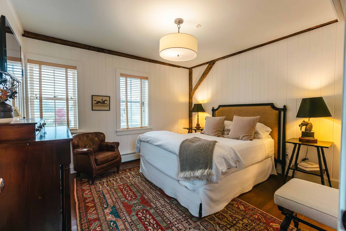 A bedroom with a large bed, two bedside tables with lamps, a wooden dresser, a leather armchair, and a patterned rug.