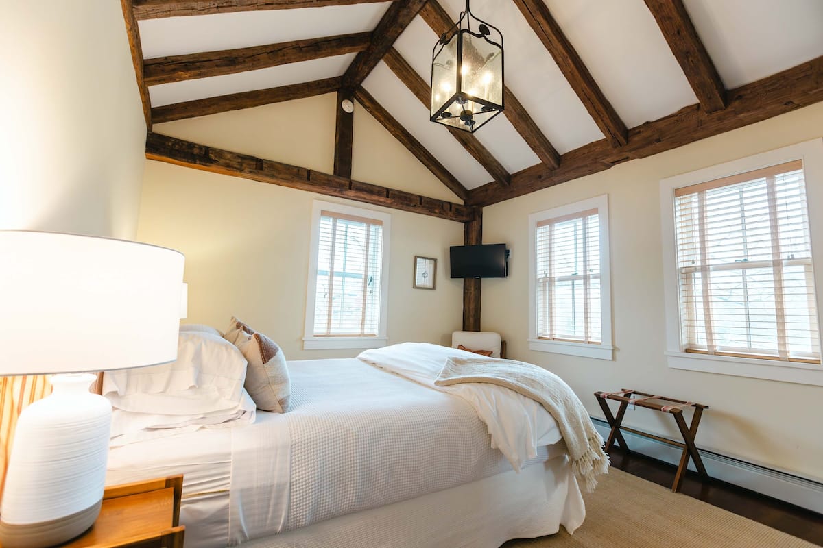 A bedroom with a large bed, wooden beams, a flat-screen TV, and a window with blinds.