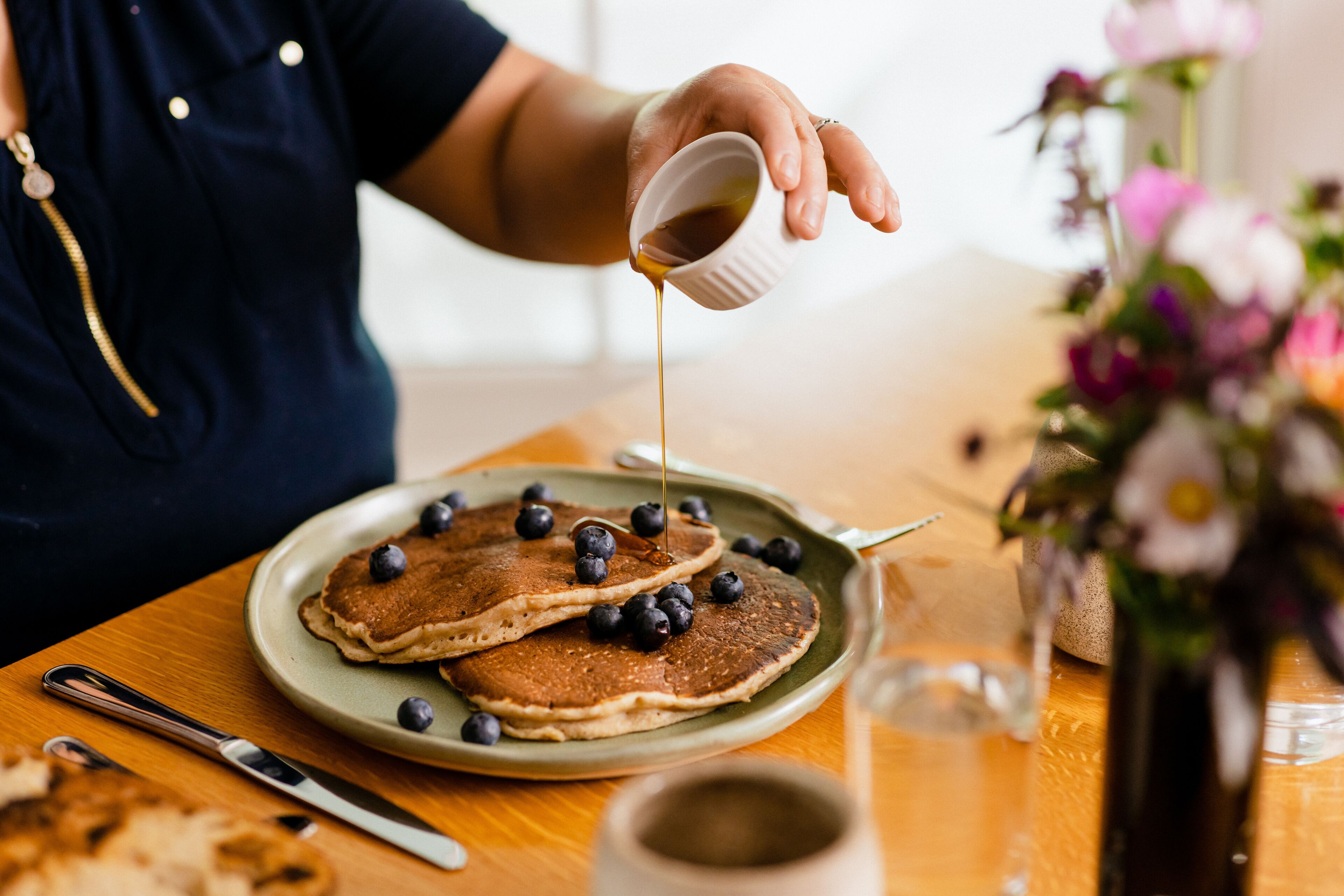 A breakfast setting with pancakes and blueberries on a plate, a cup of coffee, and a vase of flowers.