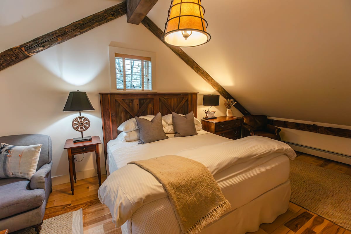 A cozy bedroom with a large bed, a wooden headboard, a nightstand with a lamp, a chair, and a window with blinds.