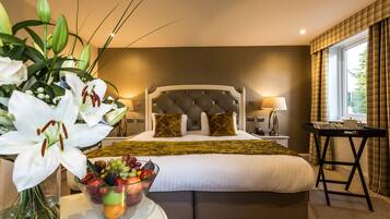 Habitación de hotel con una cama grande, una mesita de noche con frutero, una lámpara y vistas al exterior a través de una ventana.