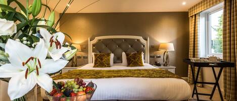 A hotel room with a large bed, a bedside table with a fruit bowl, a lamp, and a view of the outdoors through a window.