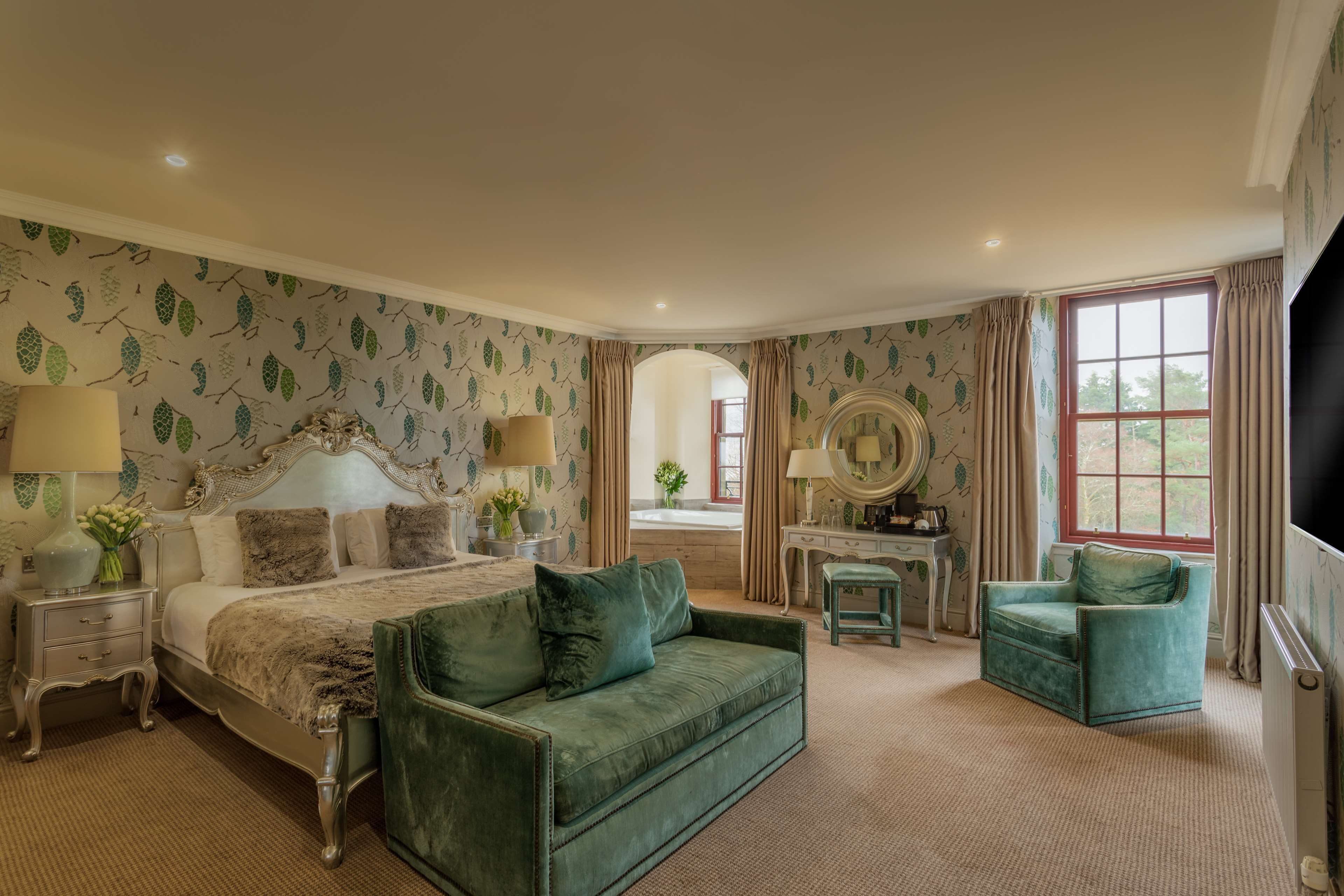 A spacious bedroom with a large bed, a green sofa, a chair, a vanity with a mirror, and a window with curtains.