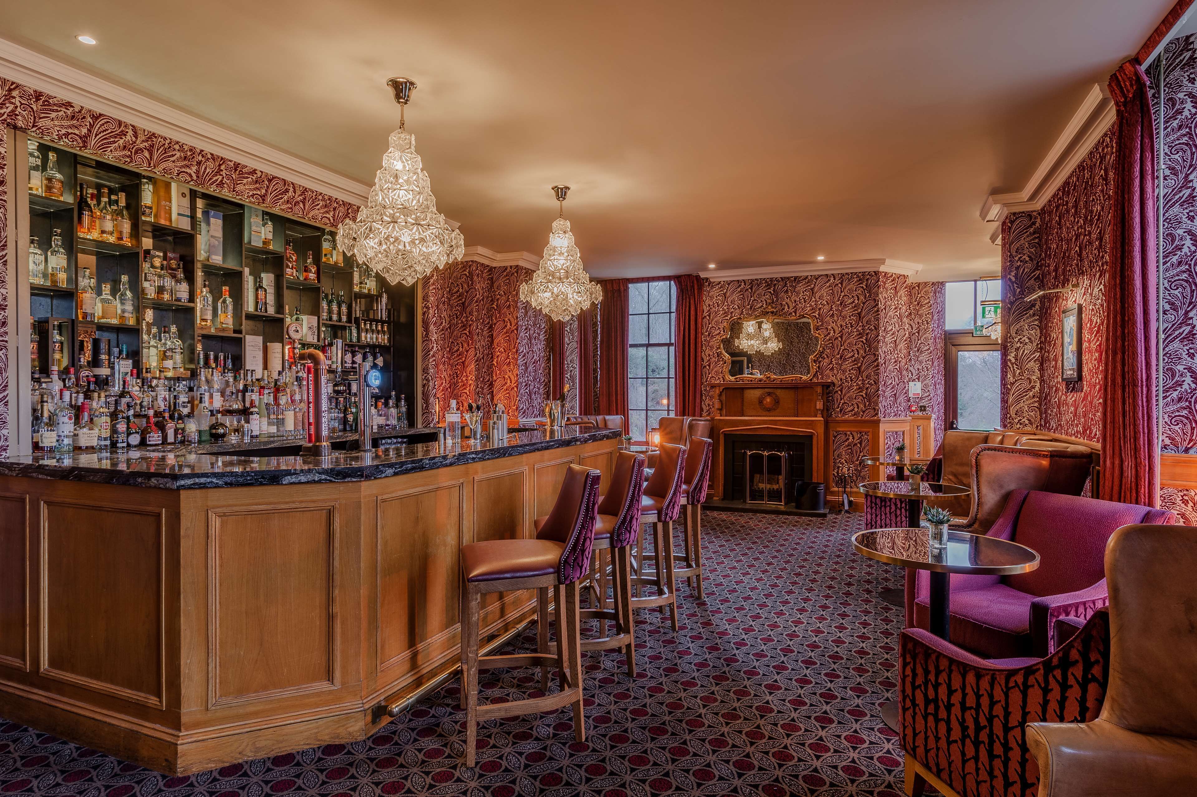 A bar area with a wooden bar counter, multiple stools, and a variety of bottles. There are two chandeliers hanging from the ceiling, and the room has patterned wallpaper and a patterned carpet.