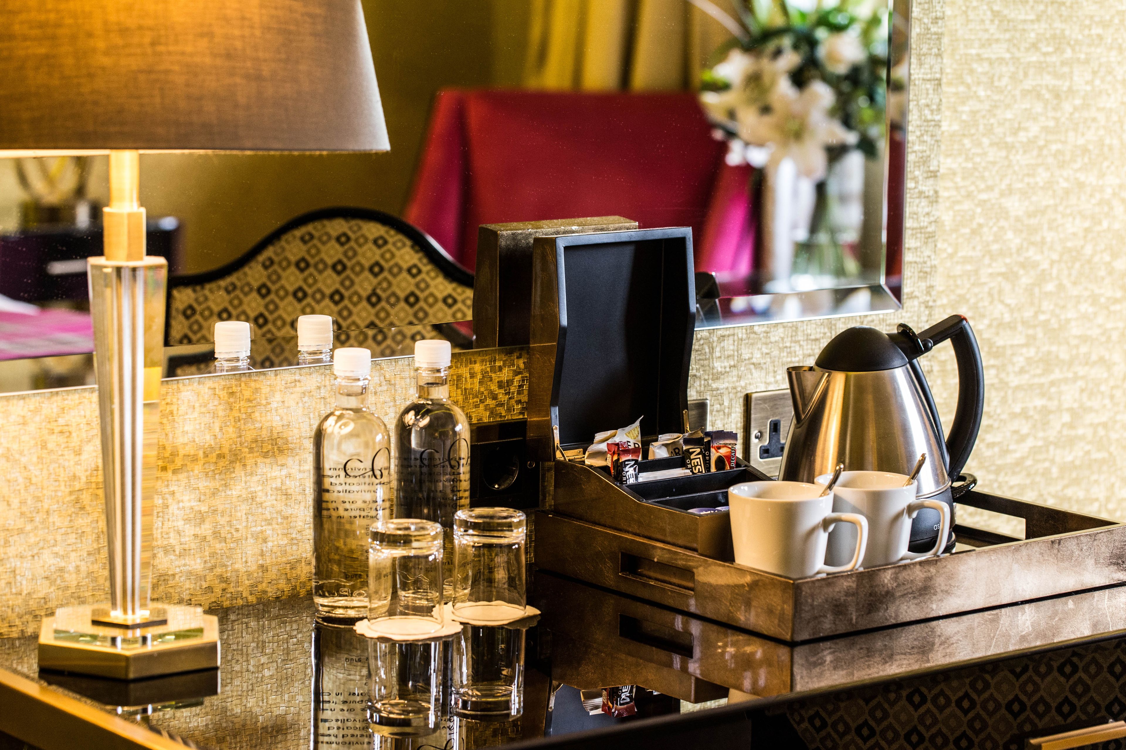 A hotel room's minibar with a lamp, bottles, glasses, and a coffee pot.
