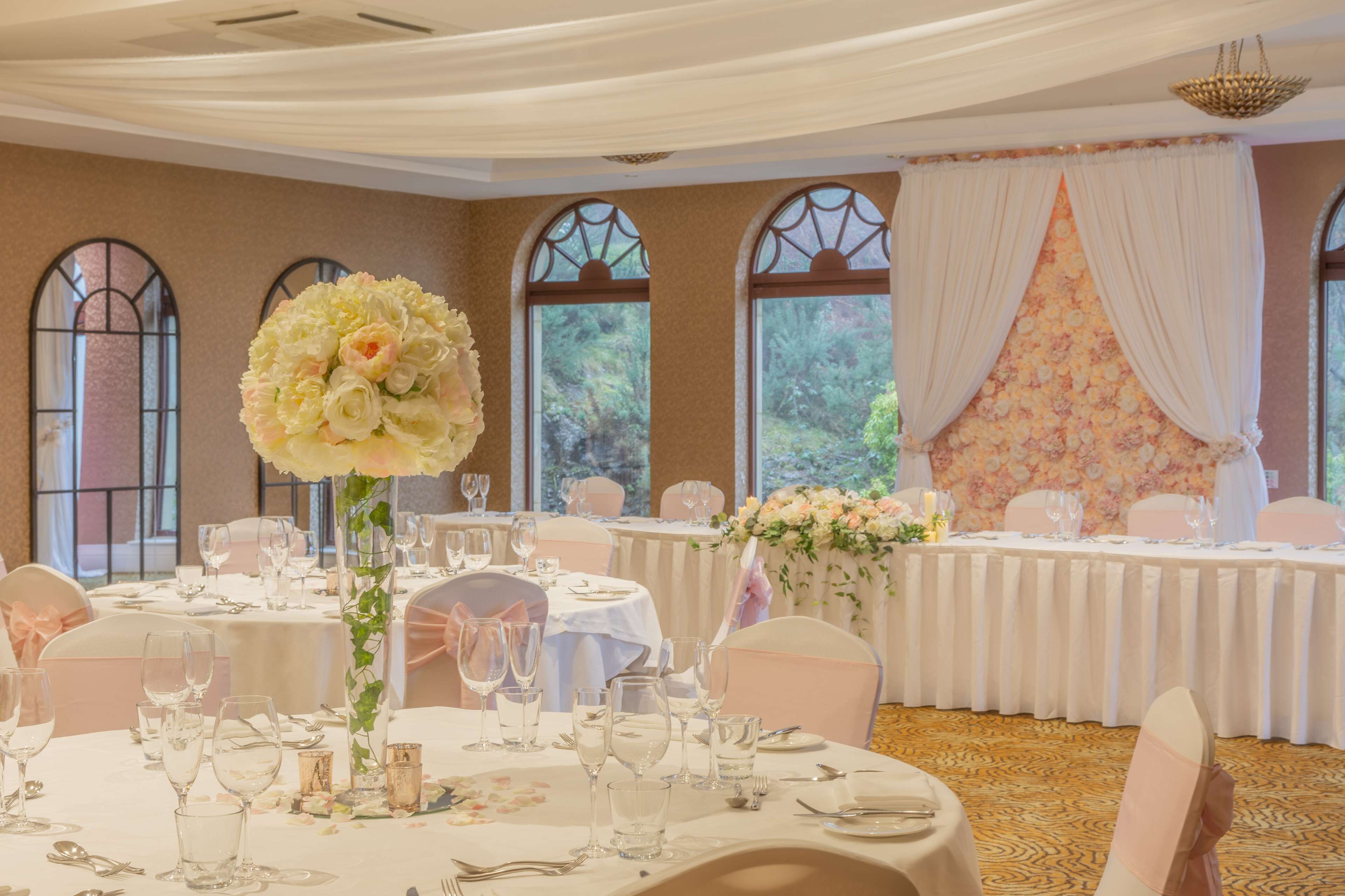 A banquet hall with round tables set for a formal event, large windows with decorative glass panes, and a floral backdrop.