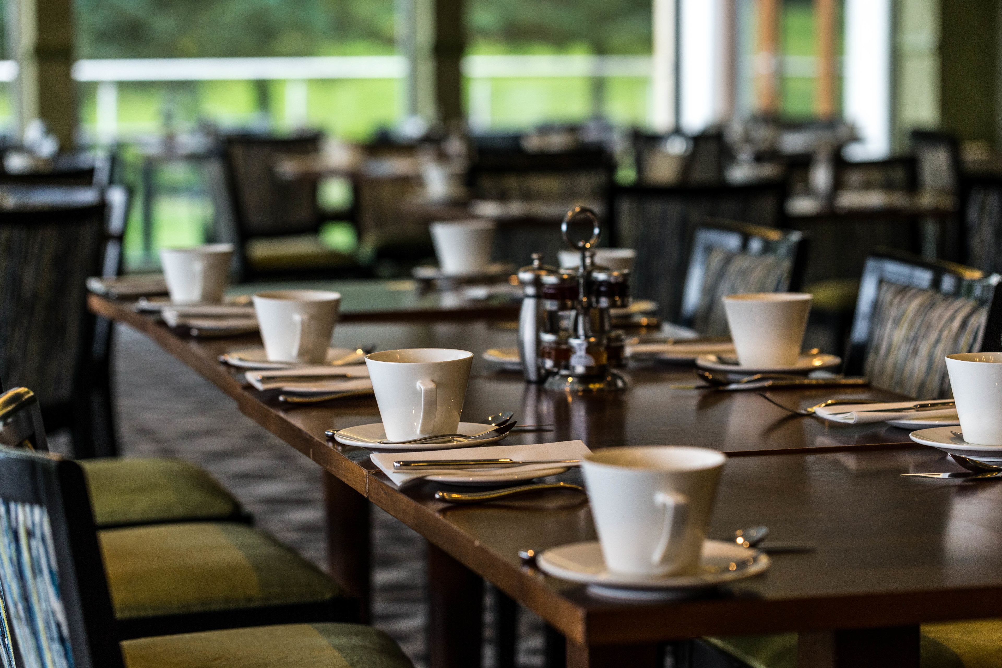 A dining table set with white cups, saucers, and cutlery, with chairs upholstered in green and black.