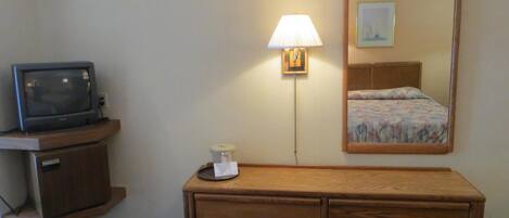 Habitación de hotel con cómoda de madera, televisor sobre una repisa, lámpara de noche y espejo que refleja una cama con colcha estampada.