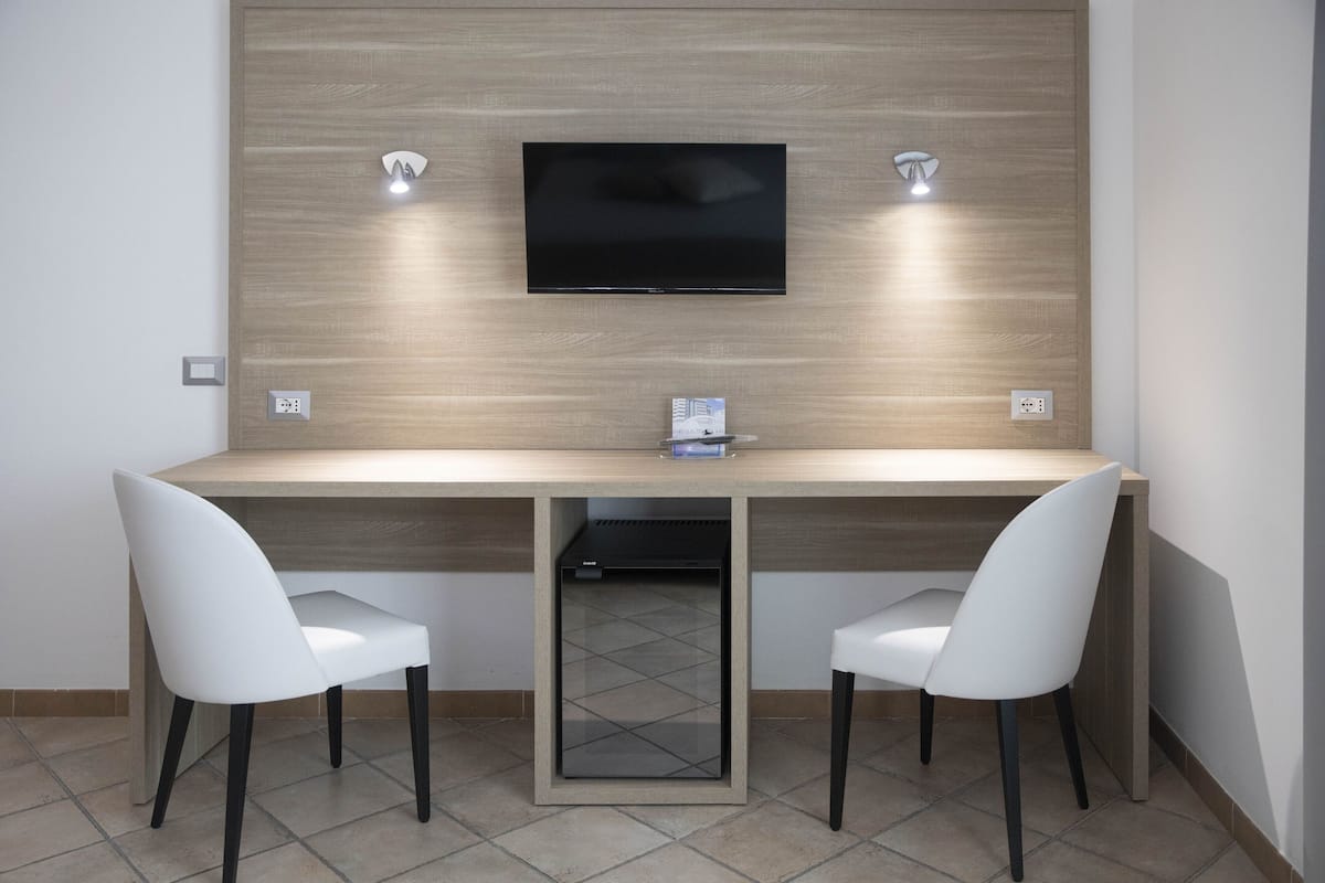 A modern room with a wooden wall, a flat-screen TV mounted above a desk, two white chairs, and a small cabinet below the desk.
