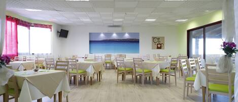 A dining area with multiple tables and chairs, a flat-screen TV mounted on the wall, and a large window with curtains.