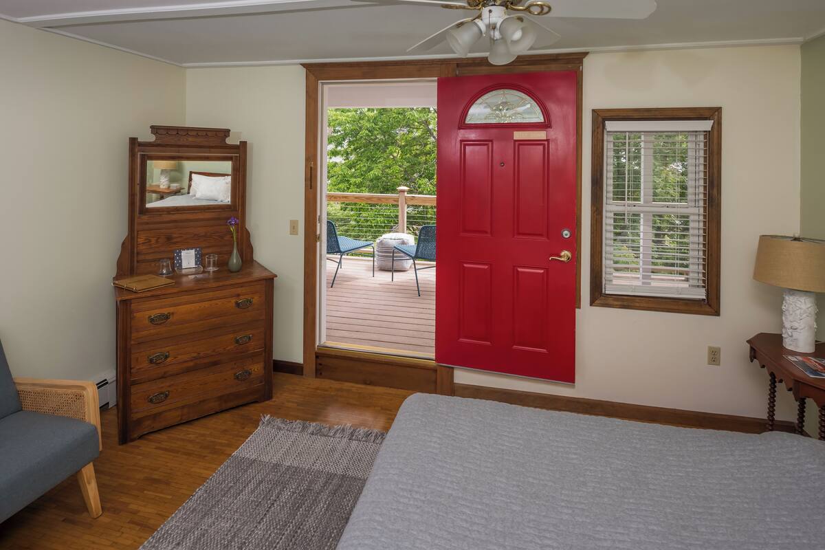 A bedroom with a wooden dresser, a red door, a window with blinds, a ceiling fan, and a view of an outdoor deck.