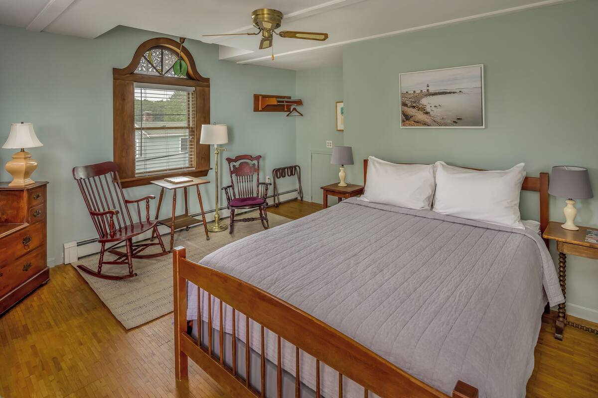 A bedroom with a large bed, wooden rocking chairs, a desk, and a window with blinds.