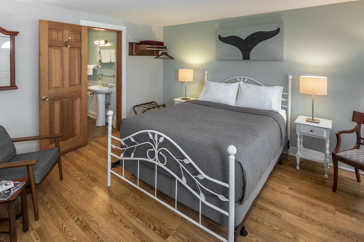 A bedroom with a metal bed frame, a bedside table with a lamp, a chair, and a wooden floor.