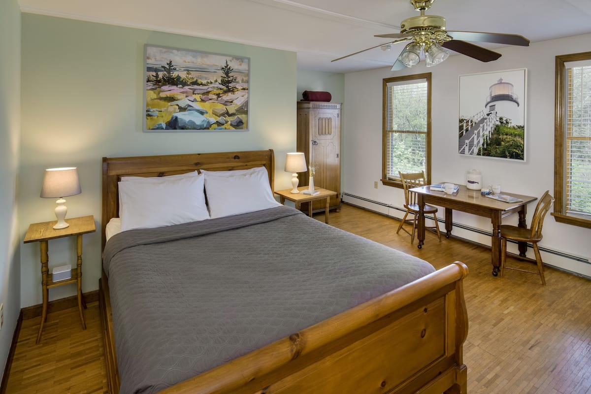 A bedroom with a wooden bed, a desk, and chairs, a ceiling fan, and artwork on the walls.