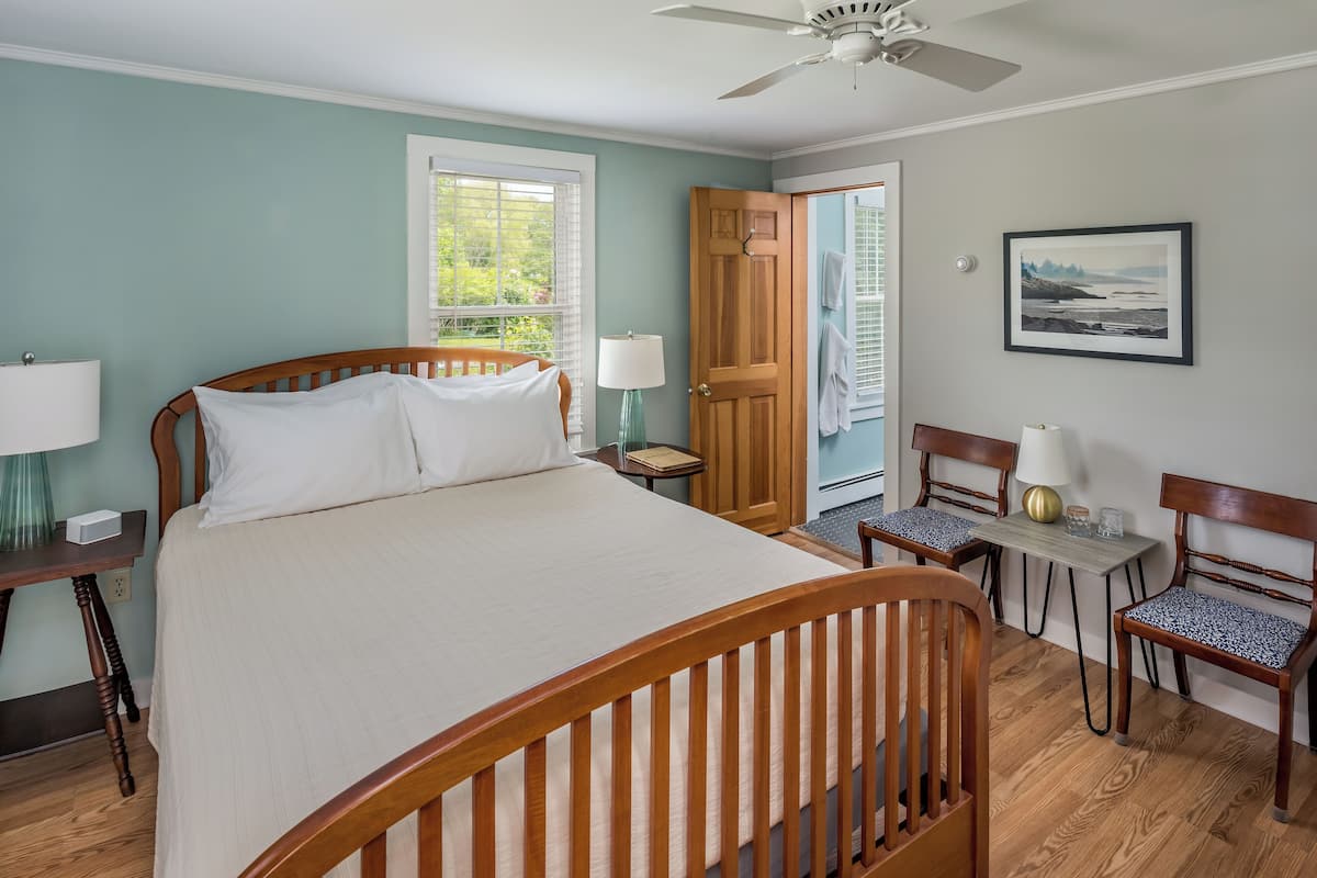 A bedroom with a wooden bed, a nightstand, a table with chairs, a ceiling fan, and a door leading to another room.