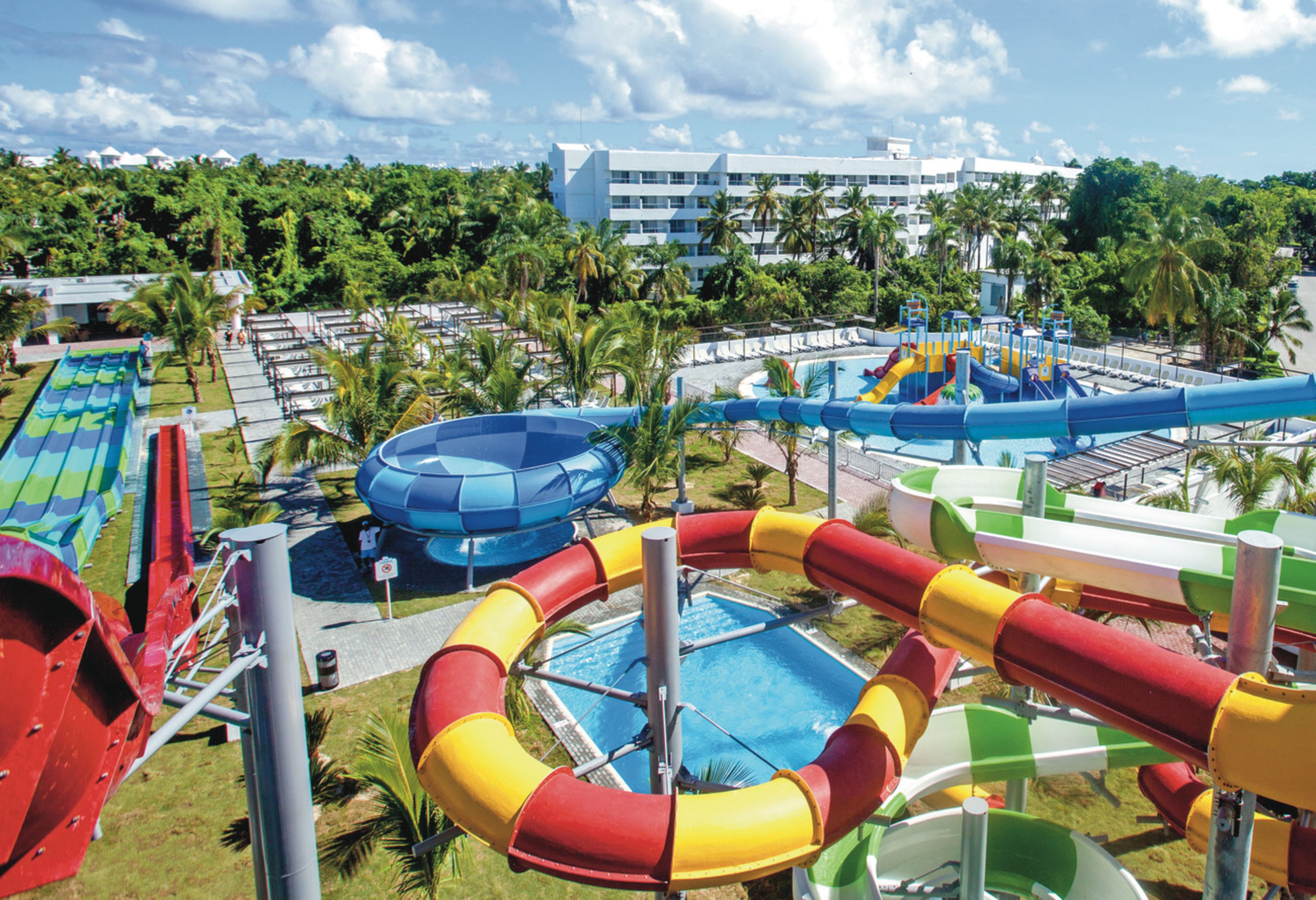 A water park with multiple slides and pools, surrounded by palm trees and a hotel in the background.