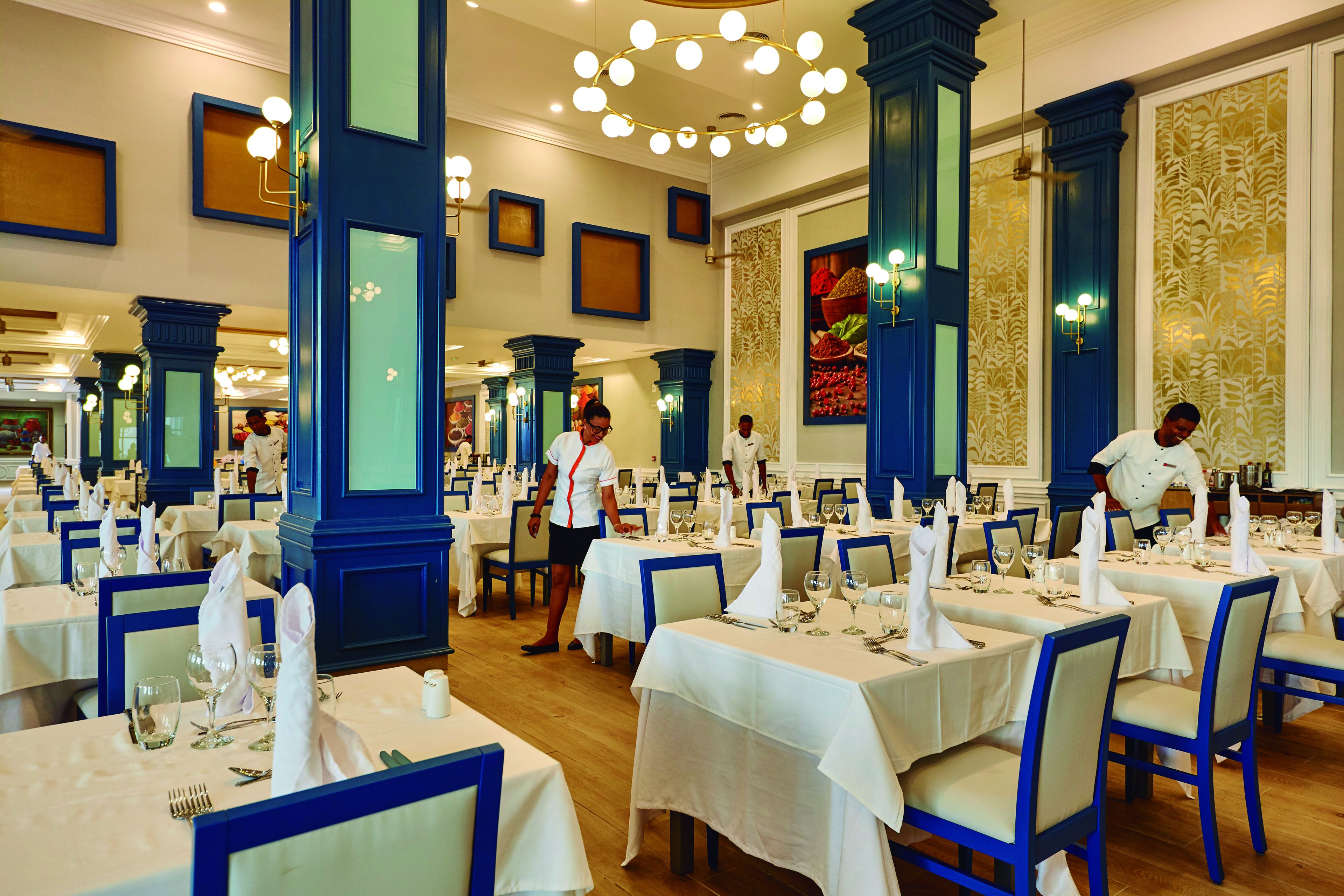 Un comedor formal con mesas dispuestas para el servicio, sillas azules y una araña.