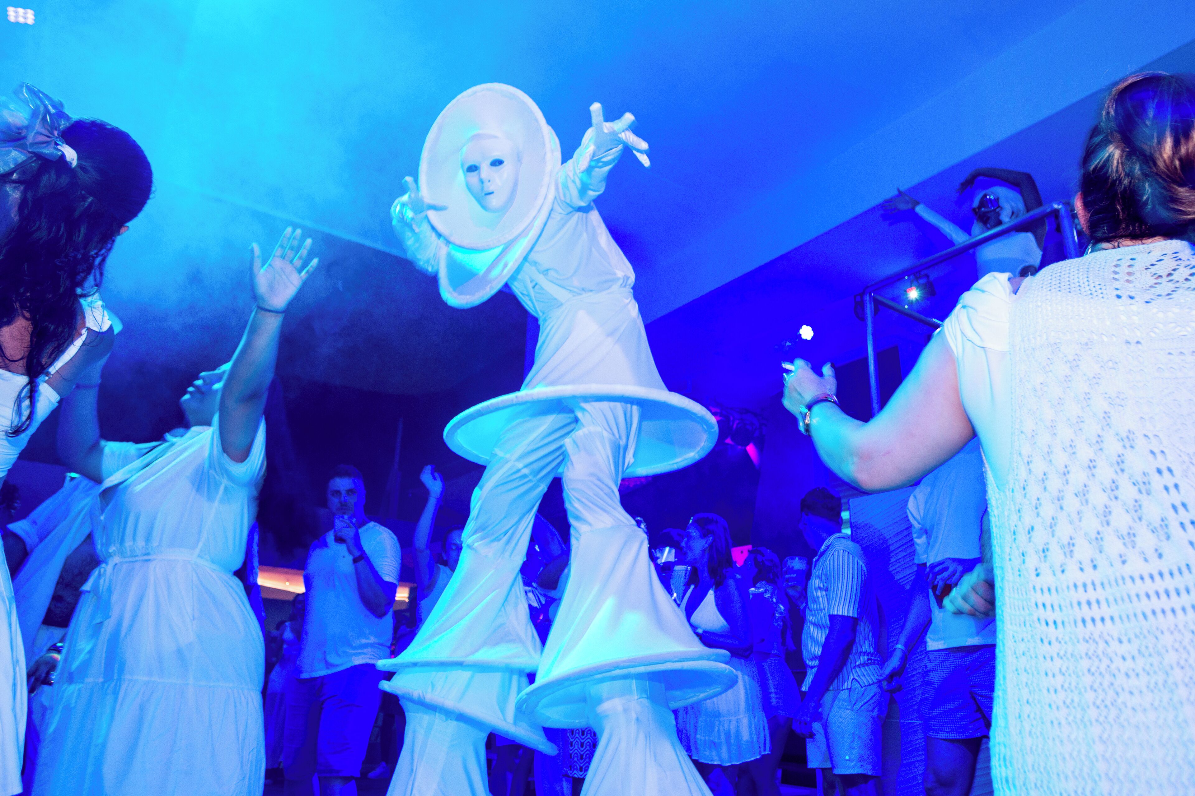 A person in a white costume with a large hat and multiple layers of fabric, surrounded by people in a dimly lit indoor setting.