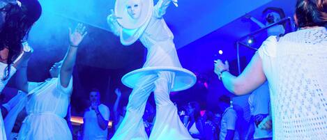A person in a white costume with a large hat and multiple layers of fabric, surrounded by people in a dimly lit indoor setting.