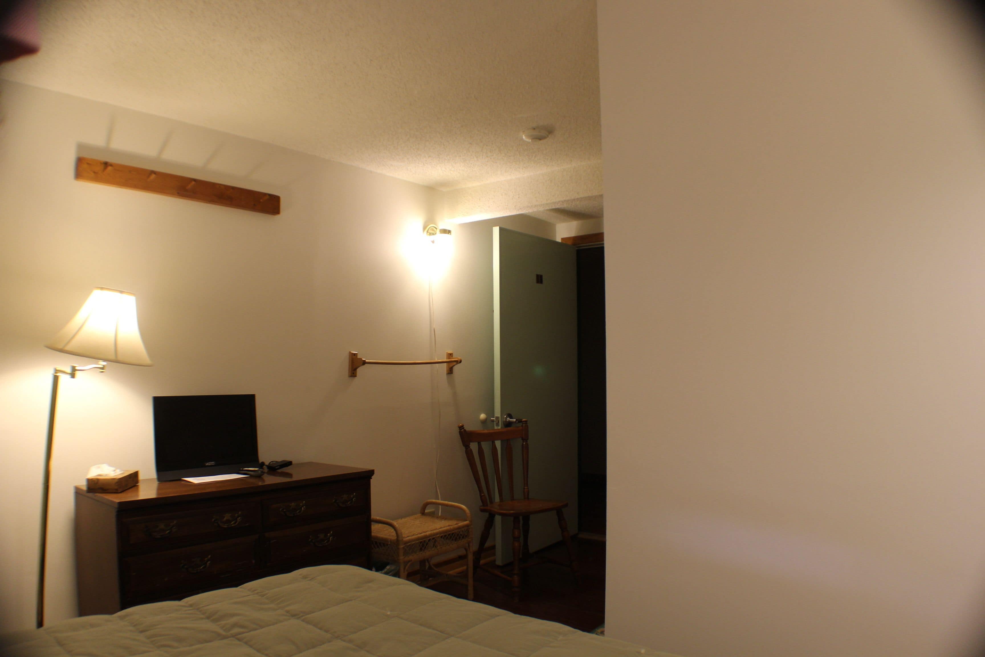 A bedroom with a bed, a dresser, a chair, a desk with a computer, and a lamp.