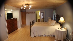 Une chambre avec un plancher en bois, un lit recouvert d’une courtepointe blanche, une table de chevet avec une lampe, une commode et un téléviseur posé sur la commode.