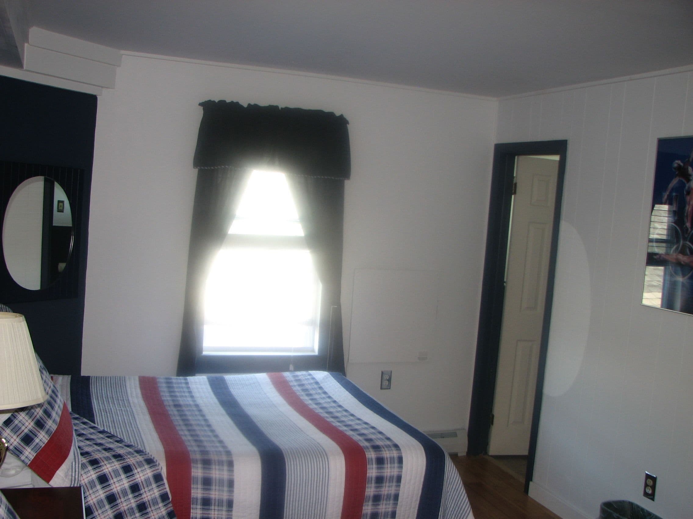 A bedroom with a bed, a window with curtains, a mirror, and a door.