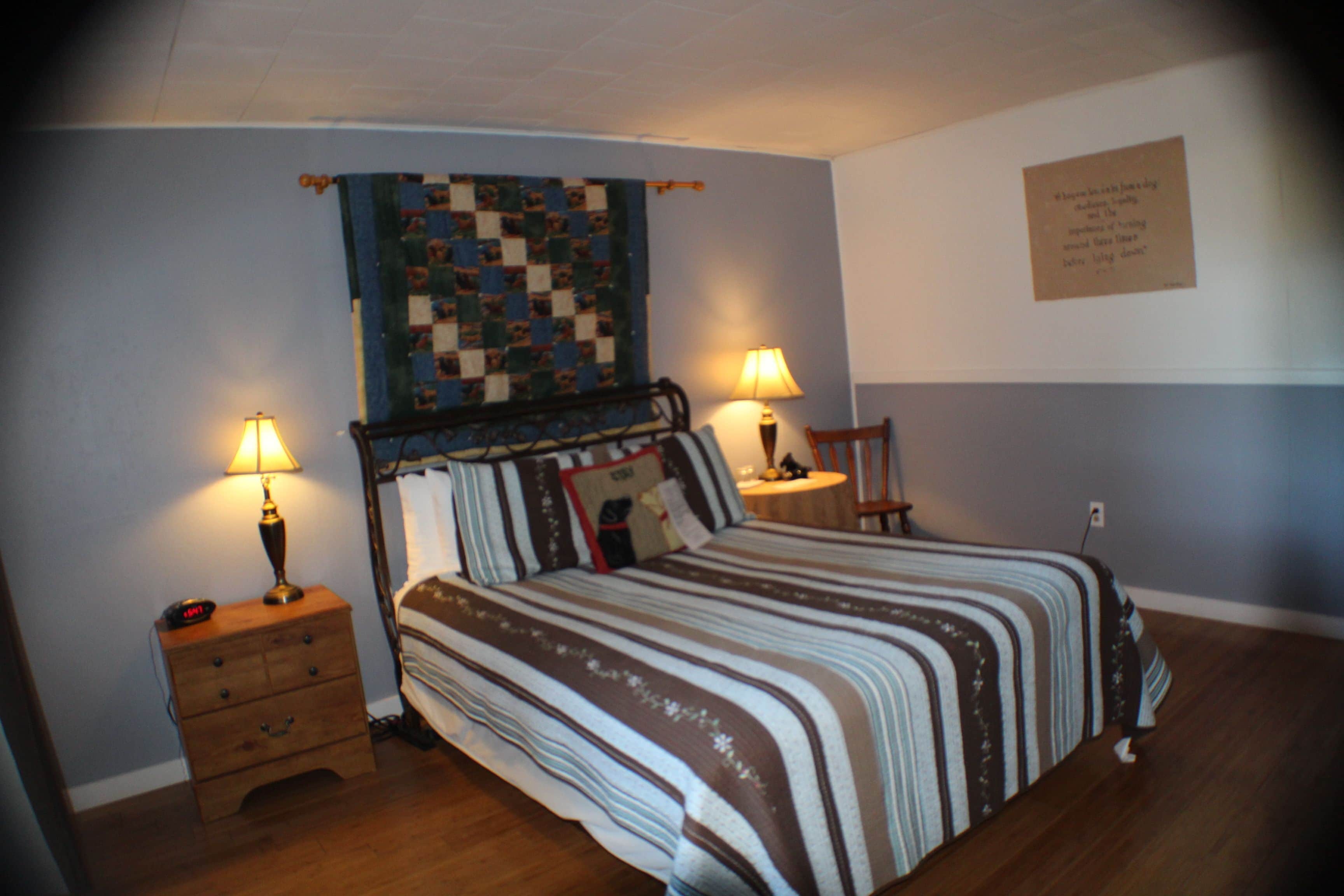 A bedroom with a bed, two bedside tables, a lamp on each table, a quilt on the wall, and a plaque on the wall.